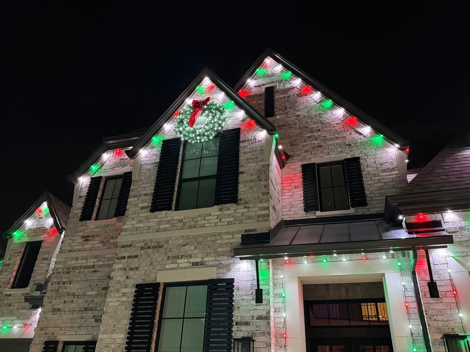 White brick two story home at night with red, green and white roofline lights and a lit wreath on the front gable.